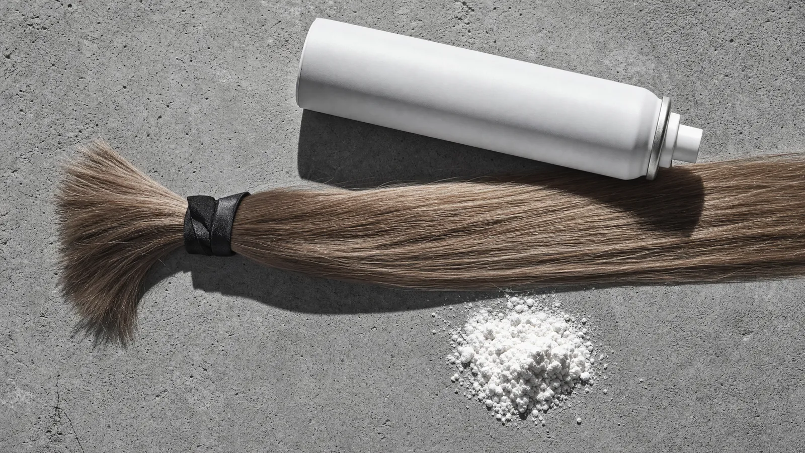 A slim ponytail of fine hair tied with black satin beside a matte white aerosol can and a heap of fine powder