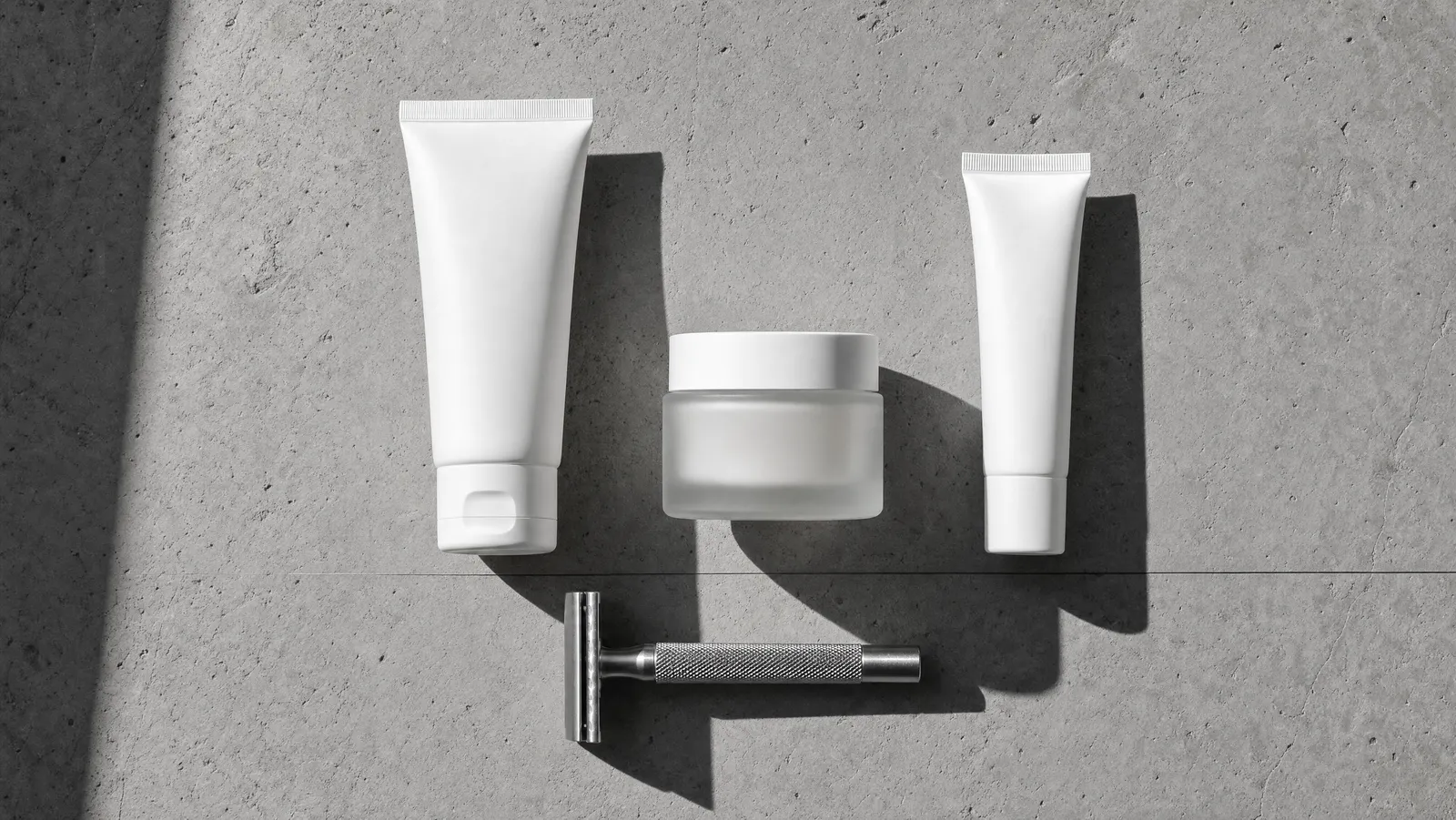 Overhead flat lay of a three-step men's skincare routine and a steel safety razor on grey concrete