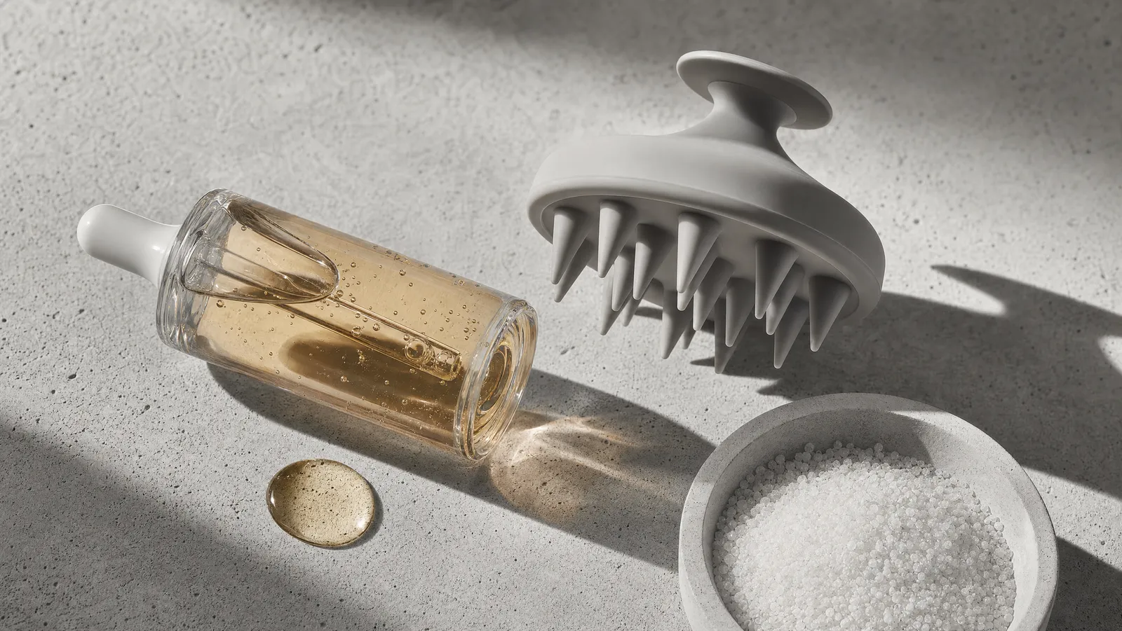 A glass dropper bottle of scalp serum, a silicone scalp brush and fine exfoliating grains on concrete